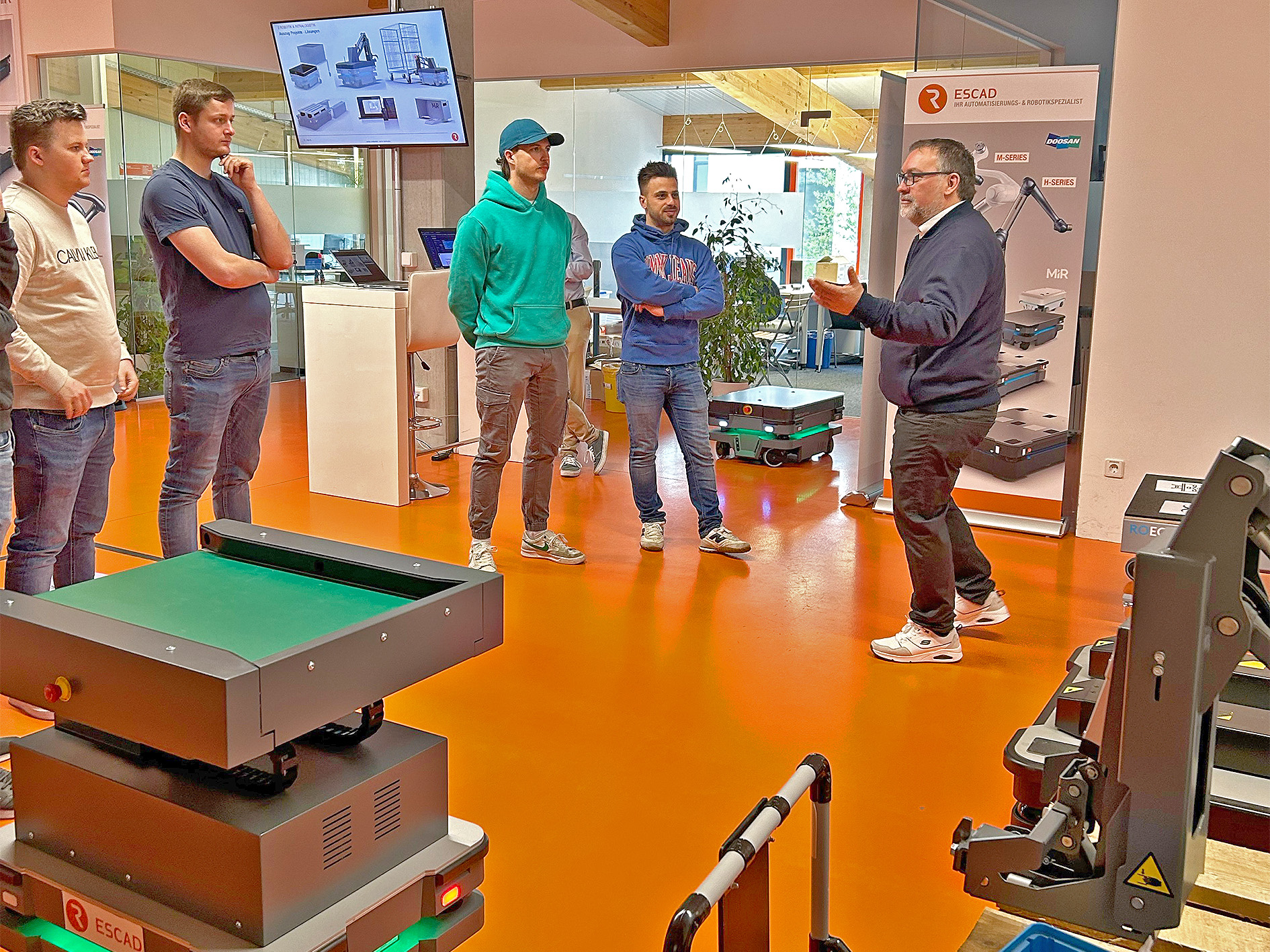 In einem Showroom stehen mehrere junge Männer im Halbkreis und begutachten verschiedene Roboter, die durch Thorsten Kulik erklärt werden.